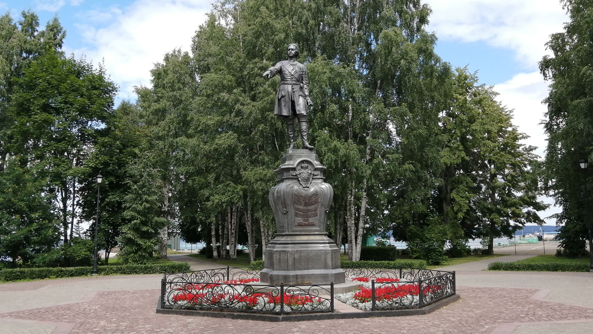 Скульптура Петра I на постаменте обрамлённом цветником и оградой на мощёной площадке в парке.