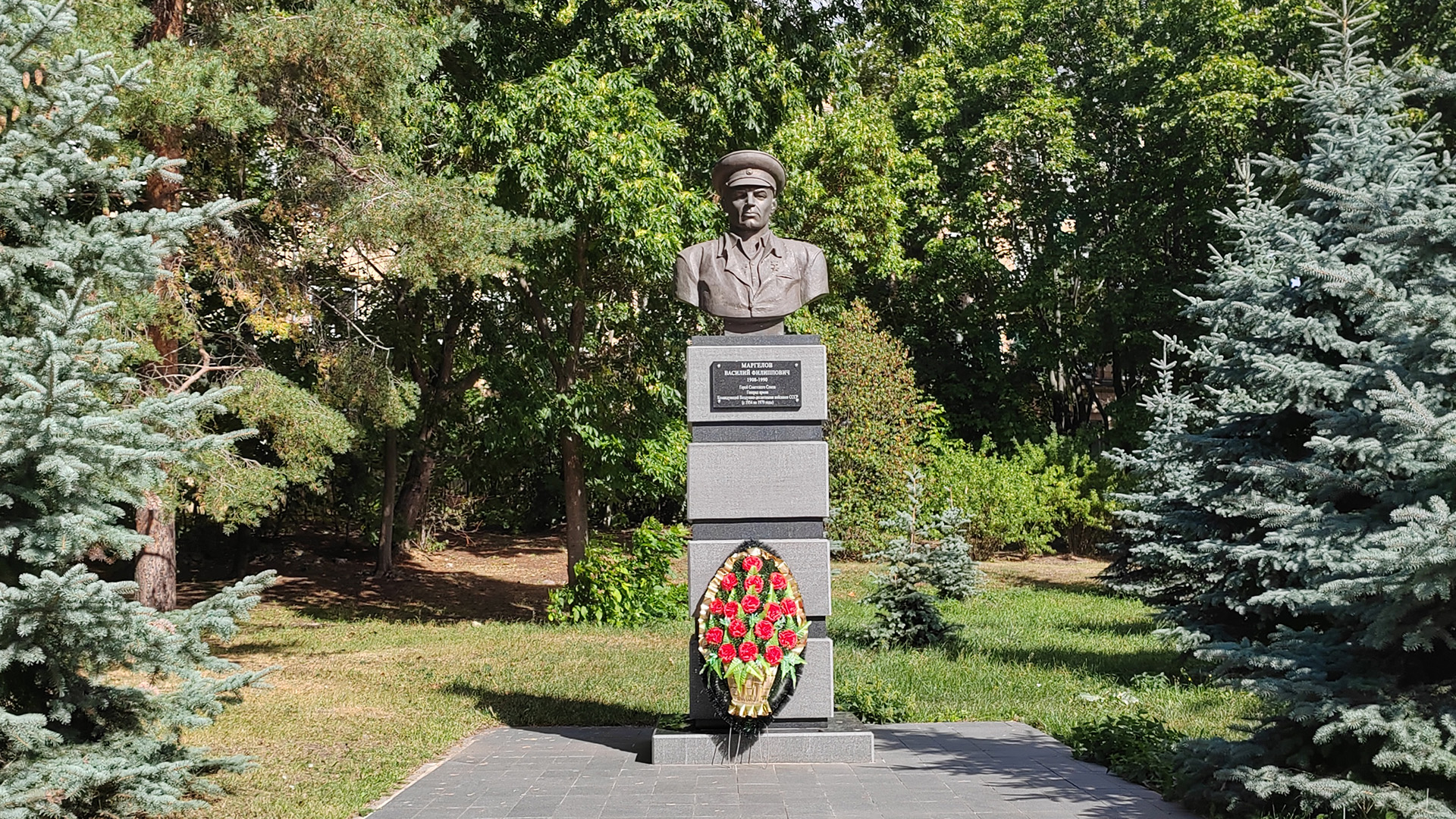 Бюст Василию Маргелову Бюст военачальника на сером каменном пьедестале расположенный среди хвойных деревьев в парке.